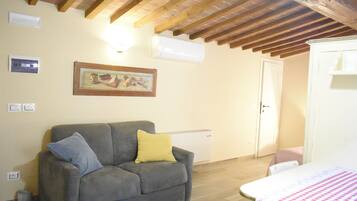 Une pièce avec un canapé gris, un lit recouvert d’une couverture à carreaux, un radiateur blanc et un plafond en bois.