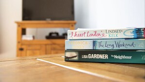 Ein Stapel Bücher auf einem Holztisch, im Hintergrund ein Fernseher.