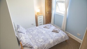 Ein kleines Schlafzimmer mit Bett, Nachttisch, Fenster mit Vorhängen und einer Tür.