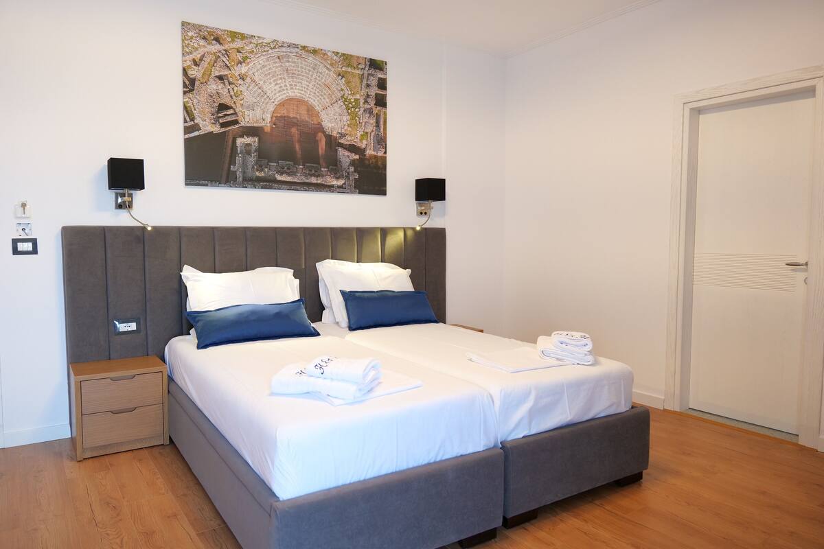 A double bed with a grey headboard, two bedside tables, a wall-mounted picture, and a white door.