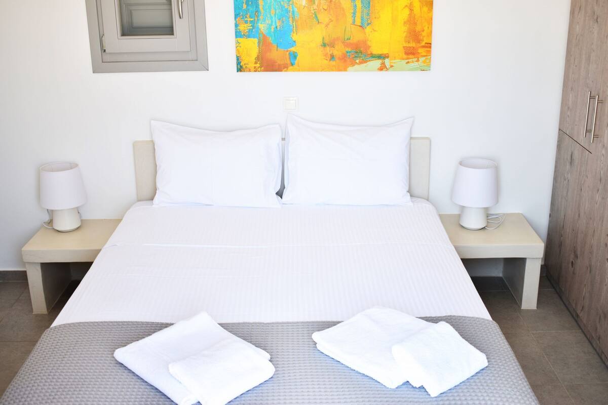 A neatly made bed with white linens, two bedside lamps, and a colorful abstract painting above the headboard.
