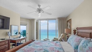 A hotel room with a large bed, a desk, a chair, a ceiling fan, and a view of the ocean.
