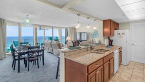 A spacious living area with a balcony overlooking the ocean, a kitchen with granite countertops, and a dining area with a table and chairs.