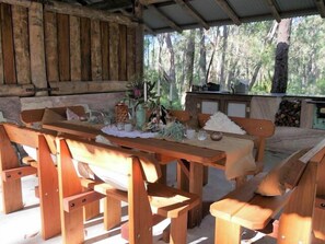 Un coin repas rustique en bois, avec des bancs et une table installés à l’extérieur, entouré d’arbres.