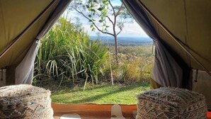 A tent with a view of a grassy landscape and a tree. Inside, there are two woven poufs and a patterned rug.