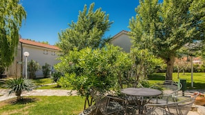 A patio area with a round table and chairs, surrounded by greenery and a building with a red roof in the background. - 1 bedroom beach front apartment in Baska (Baska)