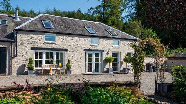 A stone house with a sloped roof, dormer windows, and a garden with various plants and a bench. - Luxury Cottage in the Scenic Borders of Scotland (Canonbie)