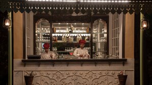 Una zona de bar con estanterías llenas de copas y botellas, dos camareros con atuendo tradicional y elaborados grabados en la pared.
