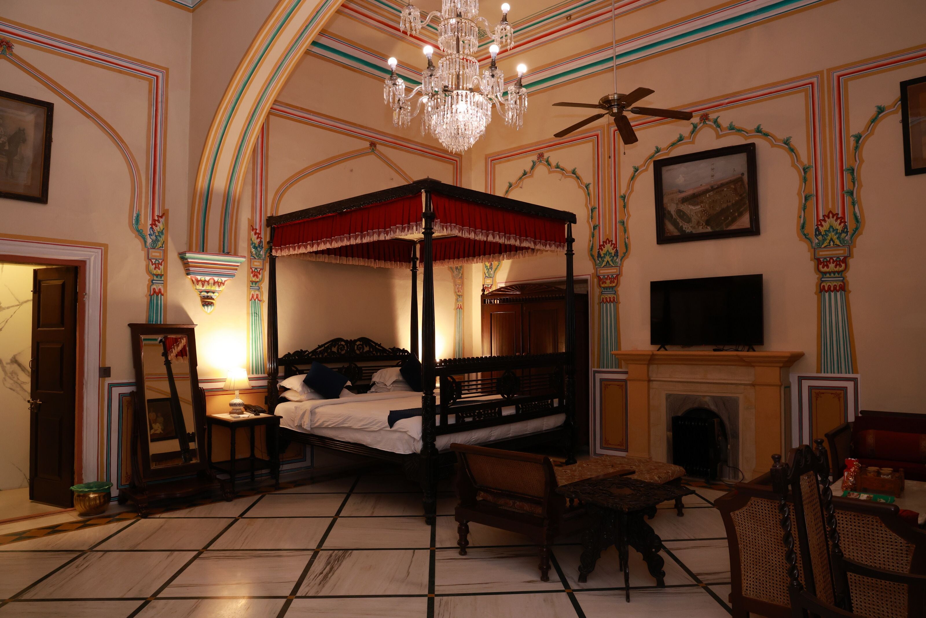 A four-poster bed, a fireplace, a television, and a chandelier in a room with ornate wall decorations.