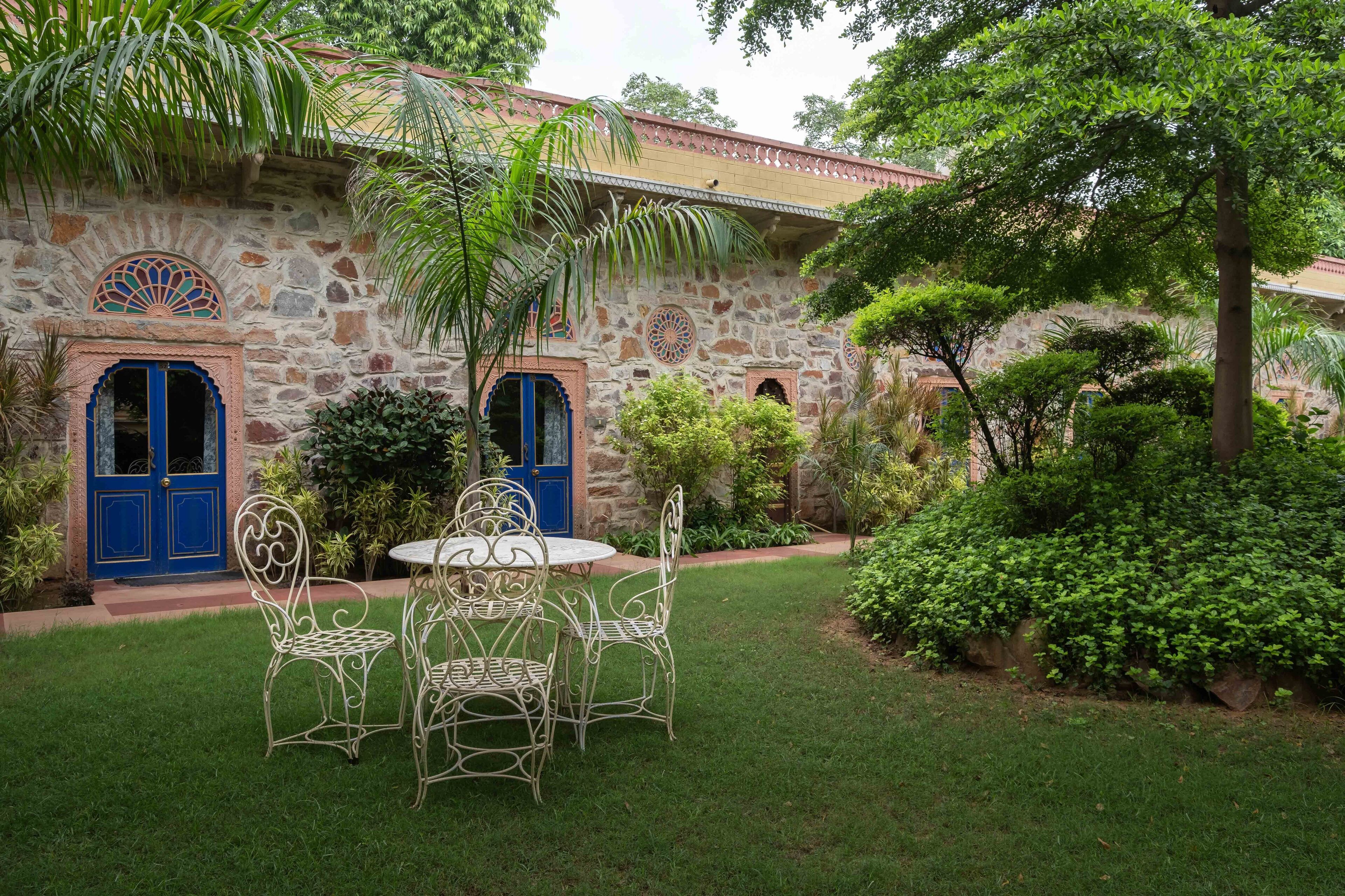 Um edifício de pedra com portas e janelas azuis, rodeado por vegetação e um pequeno jardim com cadeiras e mesa de metal.