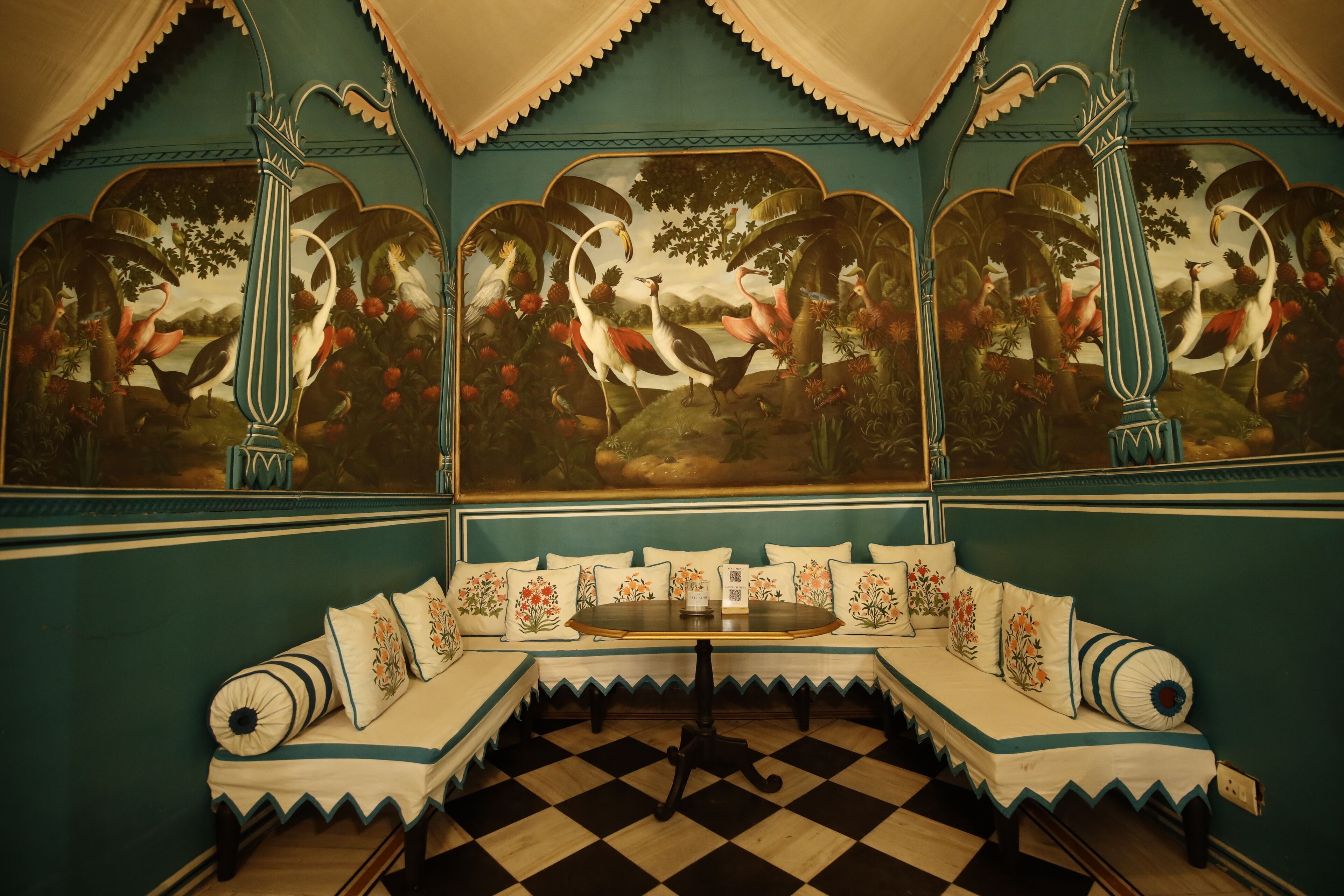 A room with a checkered floor, a sofa with floral cushions, and a painting of birds and fruits on the wall.