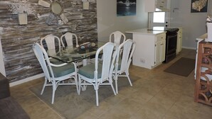 Un comedor con una mesa de vidrio y sillas de mimbre blanco, una cocina con armarios blancos y un panel de madera rústica decorativa en la pared.