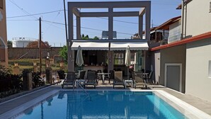 A swimming pool with lounge chairs and umbrellas, surrounded by buildings and a clear sky. - Dionisos Apartments (Corfu)