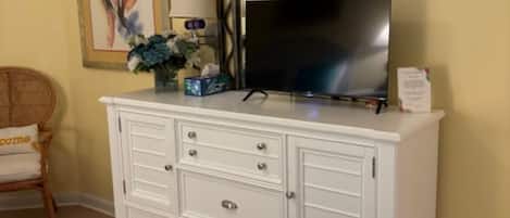 Un dormitorio con un aparador blanco, un televisor de pantalla plana, una lámpara y un cuadro en la pared.