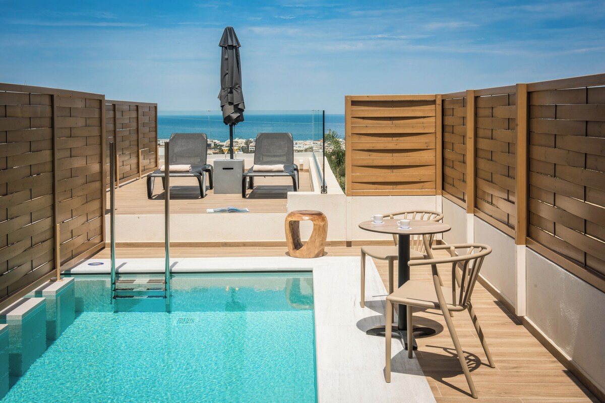 Zona de piscina con vista al mar, terraza de madera y mobiliario exterior.