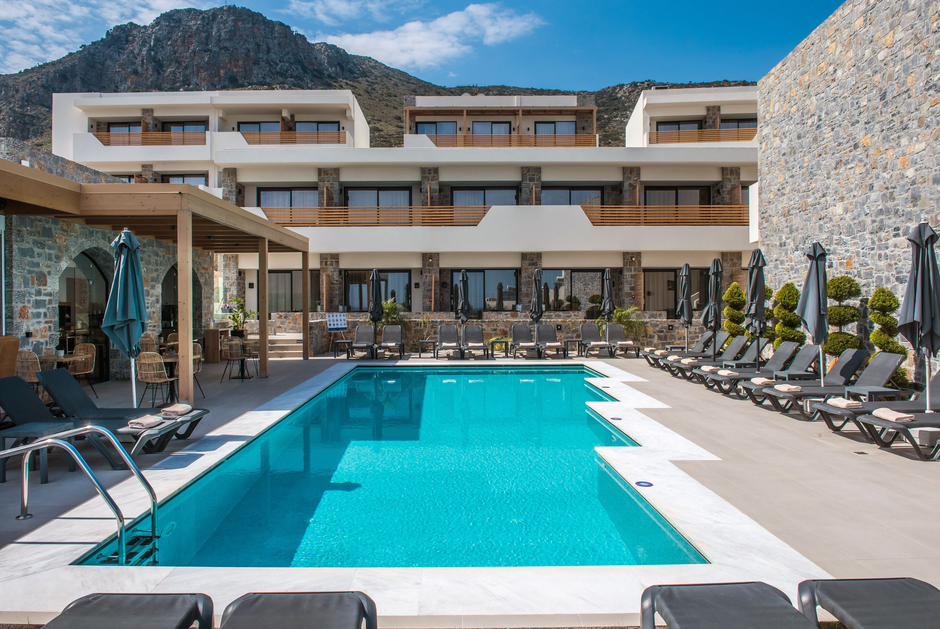 A pool area with lounge chairs and umbrellas, a stone building with multiple balconies, and a mountain backdrop.