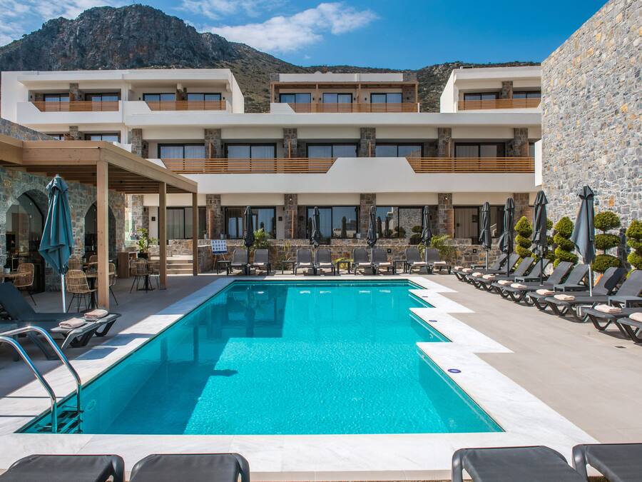 A pool area with lounge chairs and umbrellas, a stone building with multiple balconies, and a mountain backdrop.