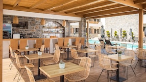 Un área de comedor al aire libre con mesas y sillas de madera, una barra con muro de piedra y una piscina al fondo.