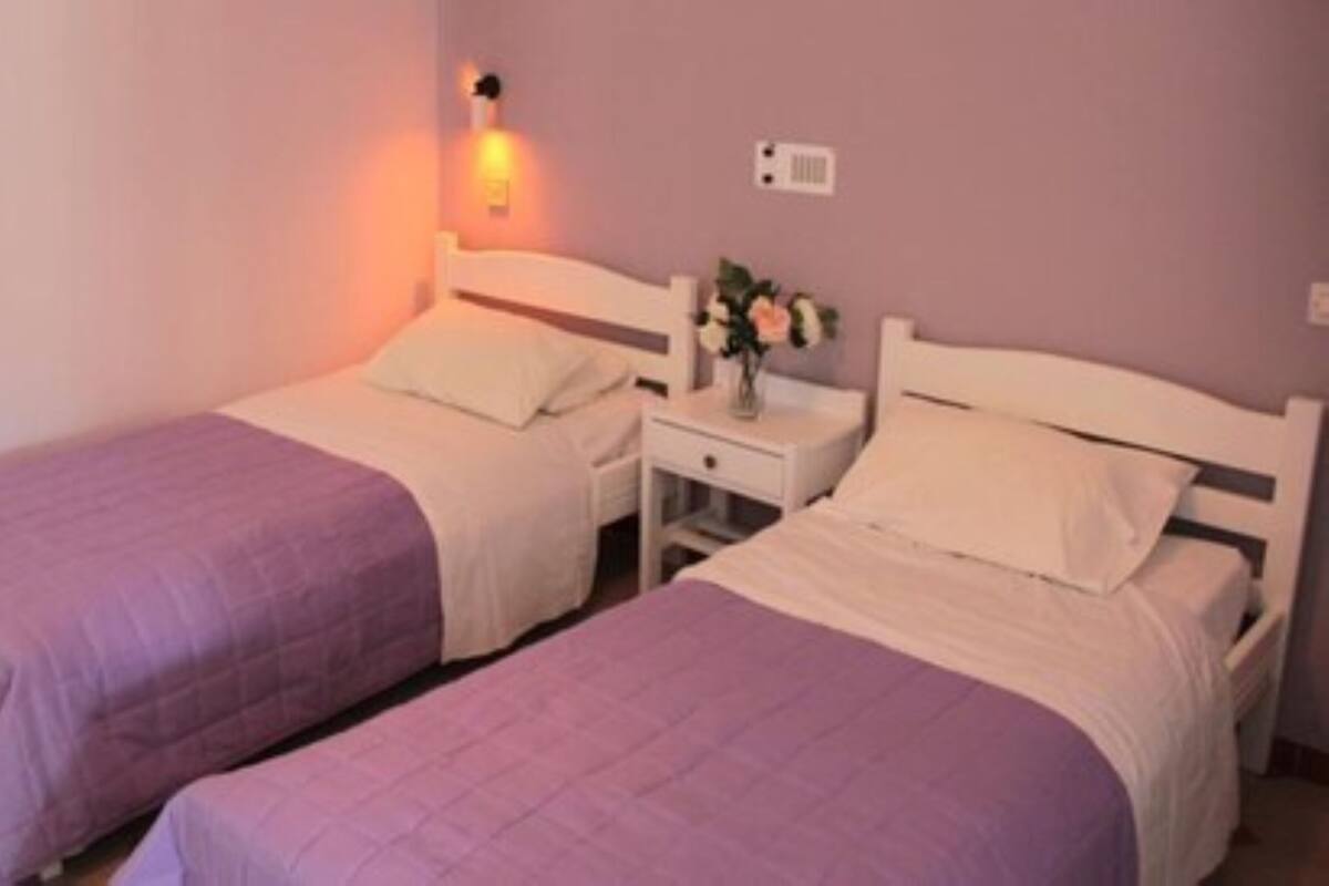 Ein Zimmer mit zwei Betten, jeweils mit weißem Kopfteil und einer violetten Tagesdecke. Dazwischen befindet sich ein Nachttisch mit einer Lampe.