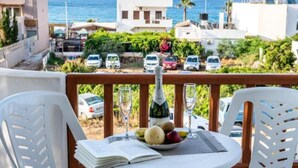 Un balcon avec des chaises et une table en plastique blanc, donnant sur une zone côtière où sont garées des voitures et où se trouvent des bâtiments.