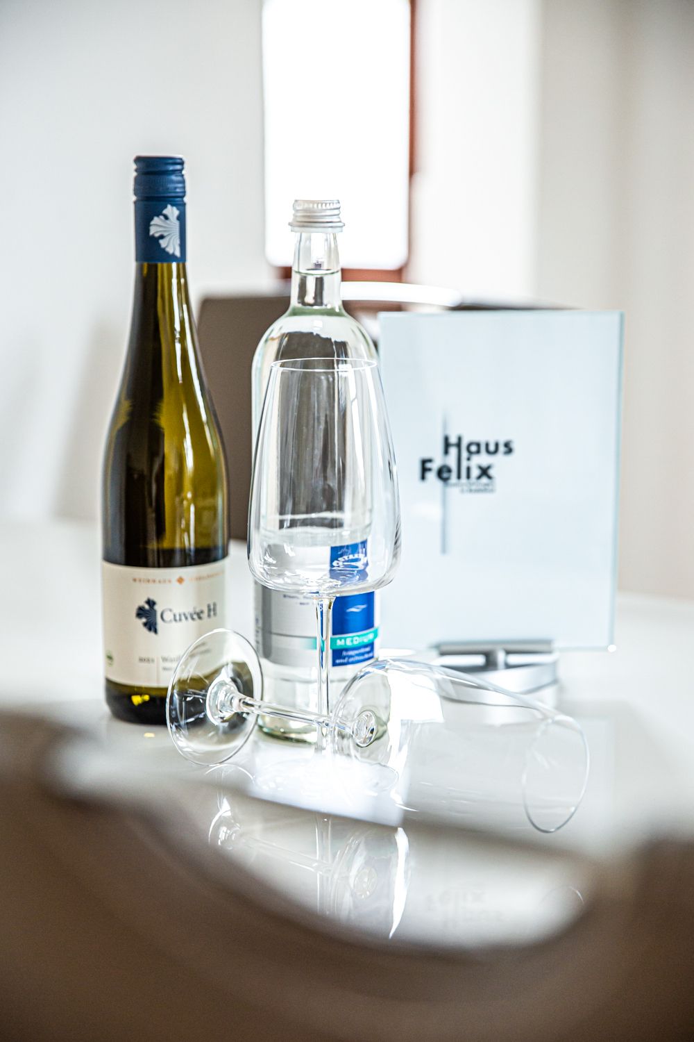 Een fles wijn, een glas en een fles water op een tafel, met een doos waarop 'Haus Felix' staat in de achtergrond.