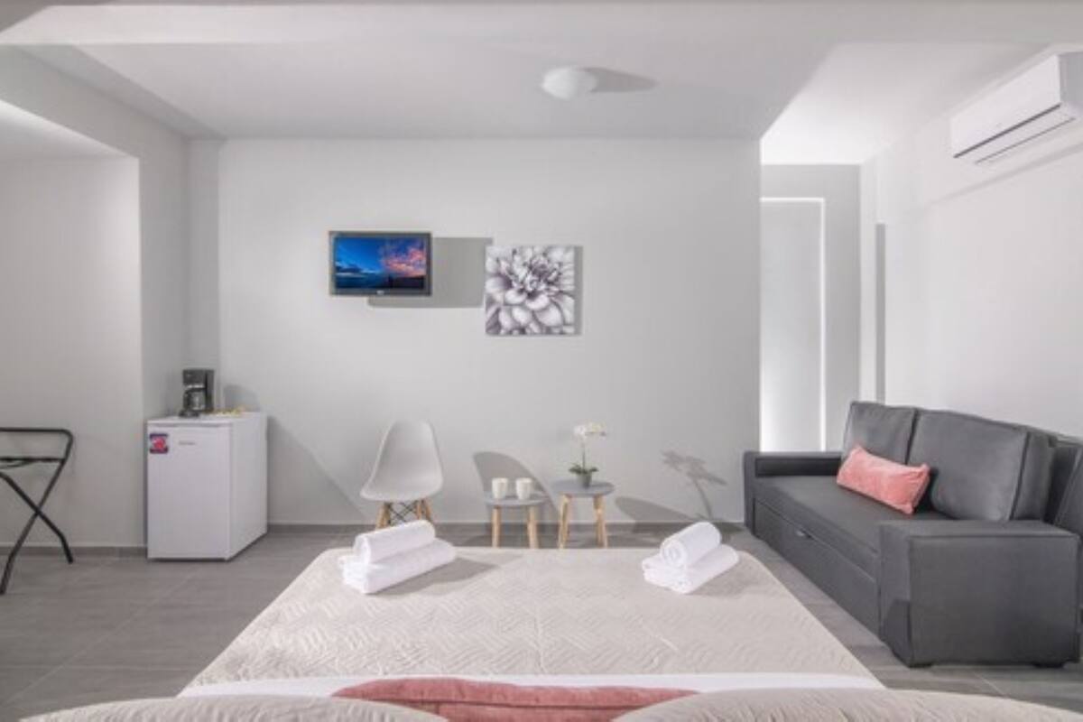 Una habitación de hotel moderna con una cama, un sofá gris, una pequeña mesa con un florero, una silla, un escritorio con una máquina de café y obras de arte en la pared.