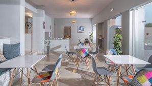 Uma área de refeições moderna, com mesas e cadeiras brancas, uma cozinha ao fundo e grandes janelas com vista.