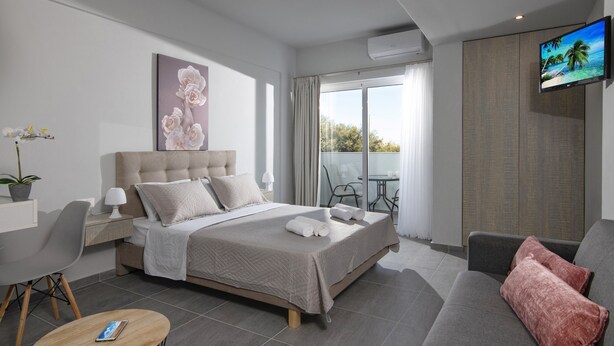 Quarto de hotel moderno com uma cama grande, TV de tela plana, varanda com vista e área de estar com sofá e poltronas.