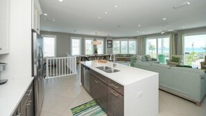 A modern kitchen with a central island, a dining area with a view, and a living room with a large sofa. - Sunset Surf 2201 Ave C 6 Bedroom Home by RedAwning (Bradenton Beach)