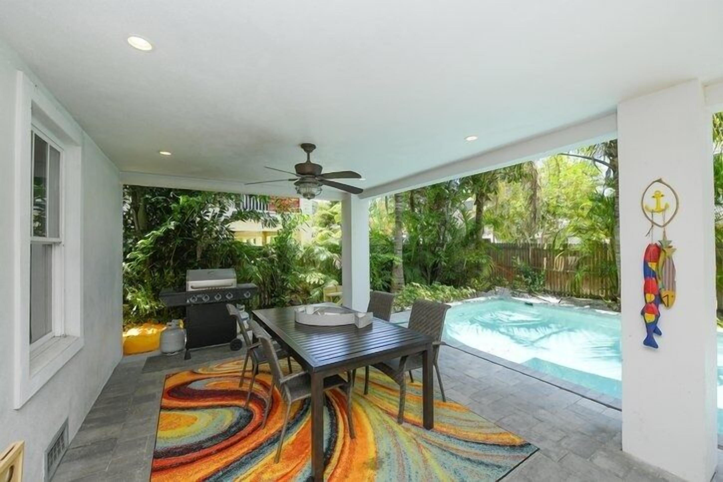 An outdoor patio with a dining table, chairs, and a colorful rug. There is a grill and a pool in the background.