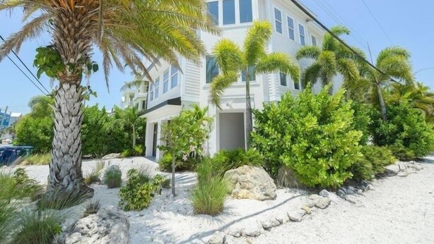 A two-story building with a white exterior, surrounded by tropical vegetation and a sandy landscape. - Sunset Surf 2201 Ave C 6 Bedroom Home by RedAwning (Bradenton Beach)