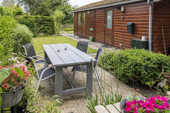 A wooden cabin with a patio area featuring a table and chairs, surrounded by well-maintained gardens and hedges.