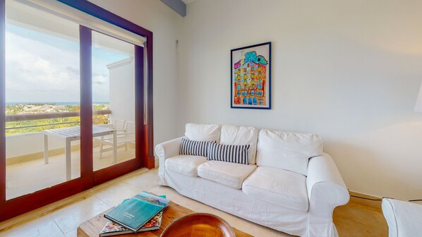 Um sofá branco com almofadas listradas, uma mesa de centro de madeira com livros e uma tigela, uma obra de arte colorida emoldurada na parede e uma varanda com mesa e cadeiras.