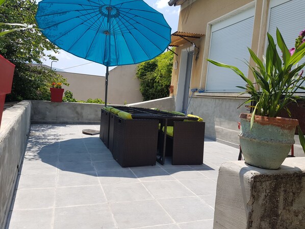 Una terraza en la azotea con un paraguas azul, mobiliario de mimbre, plantas en macetas y vistas de los edificios circundantes.