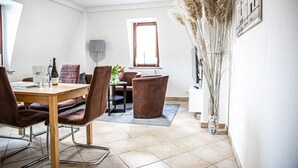 A dining area with a wooden table, brown upholstered chairs, a small side table with a vase of flowers, and a window with a view of buildings. - House Felix Weinbergblick (Radebeul)