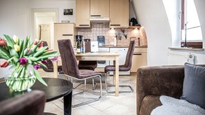 A modern kitchen with wooden cabinets, a dining table with chairs, and a view of the living area with a couch and a vase of flowers. - House Felix Weinbergblick (Radebeul)