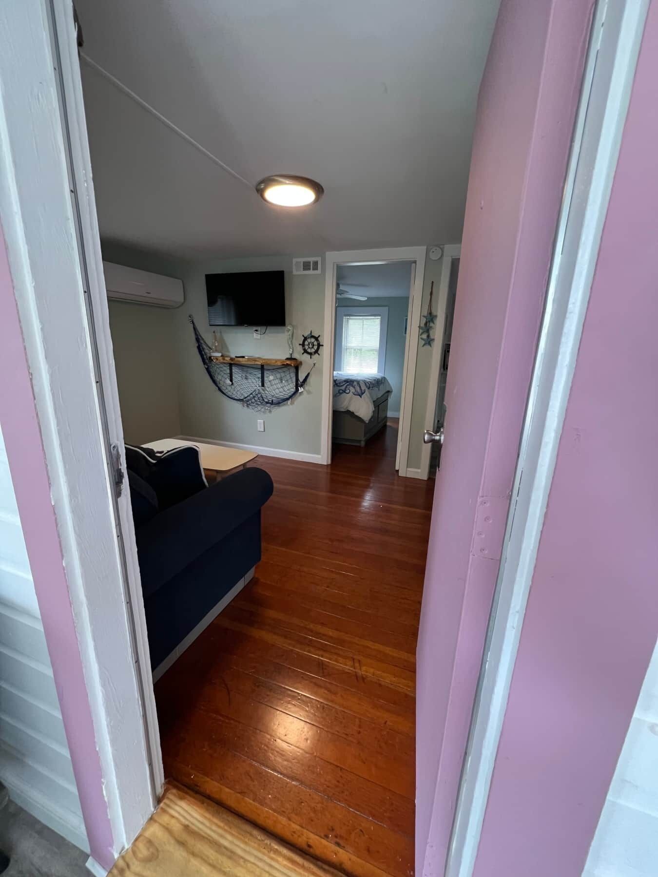 Un pasillo con piso de madera que conduce a una habitación con un sofá azul oscuro y un televisor montado en la pared.