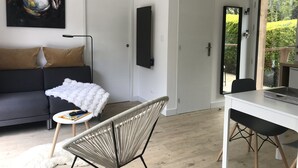 Una sala moderna con un sofá, un plaid blanco y mullido, una silla, una mesa de comedor y un ventanal con vistas al verdor.