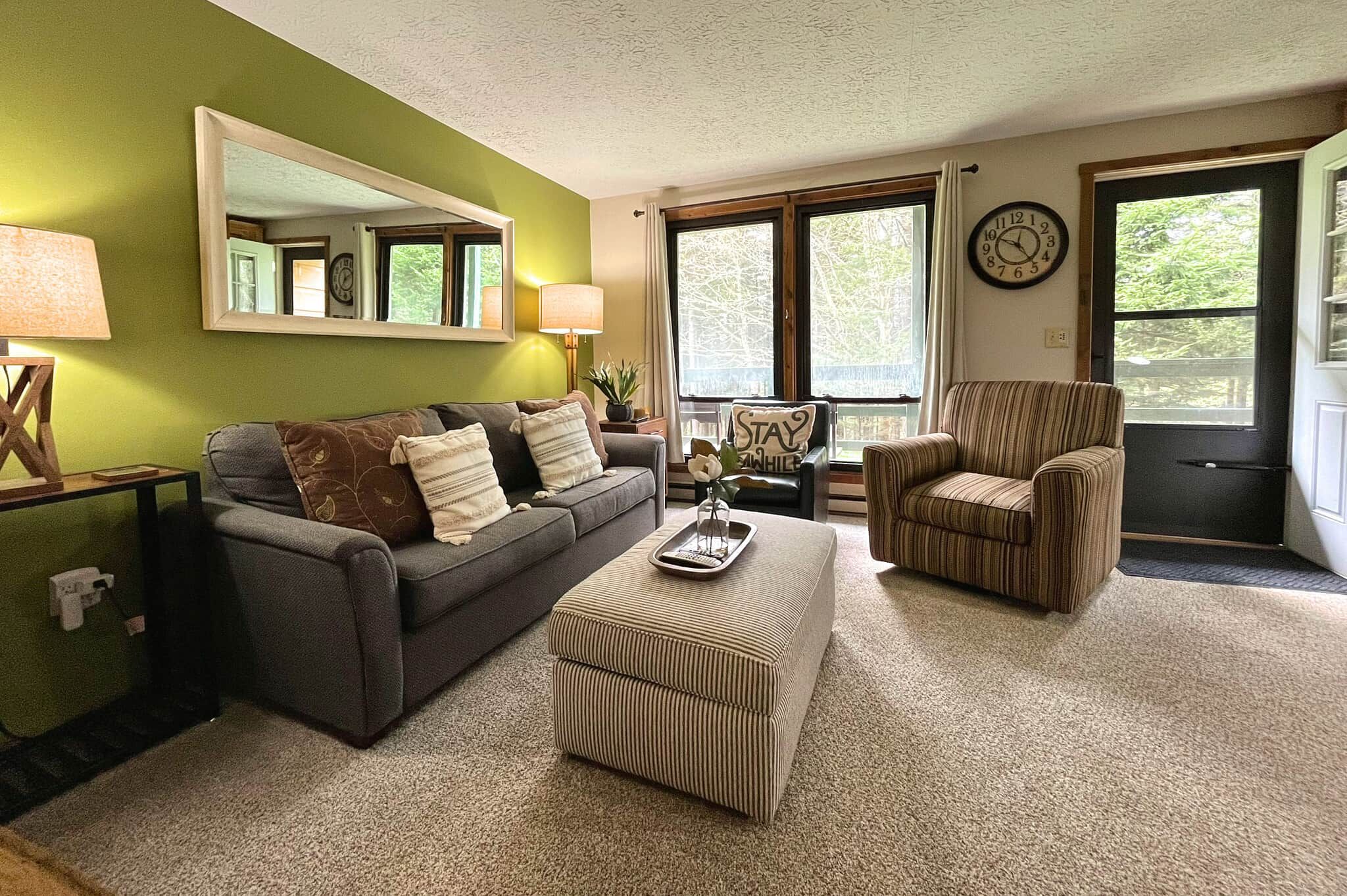 A living room with a sofa, armchair, ottoman, and a clock on the wall.