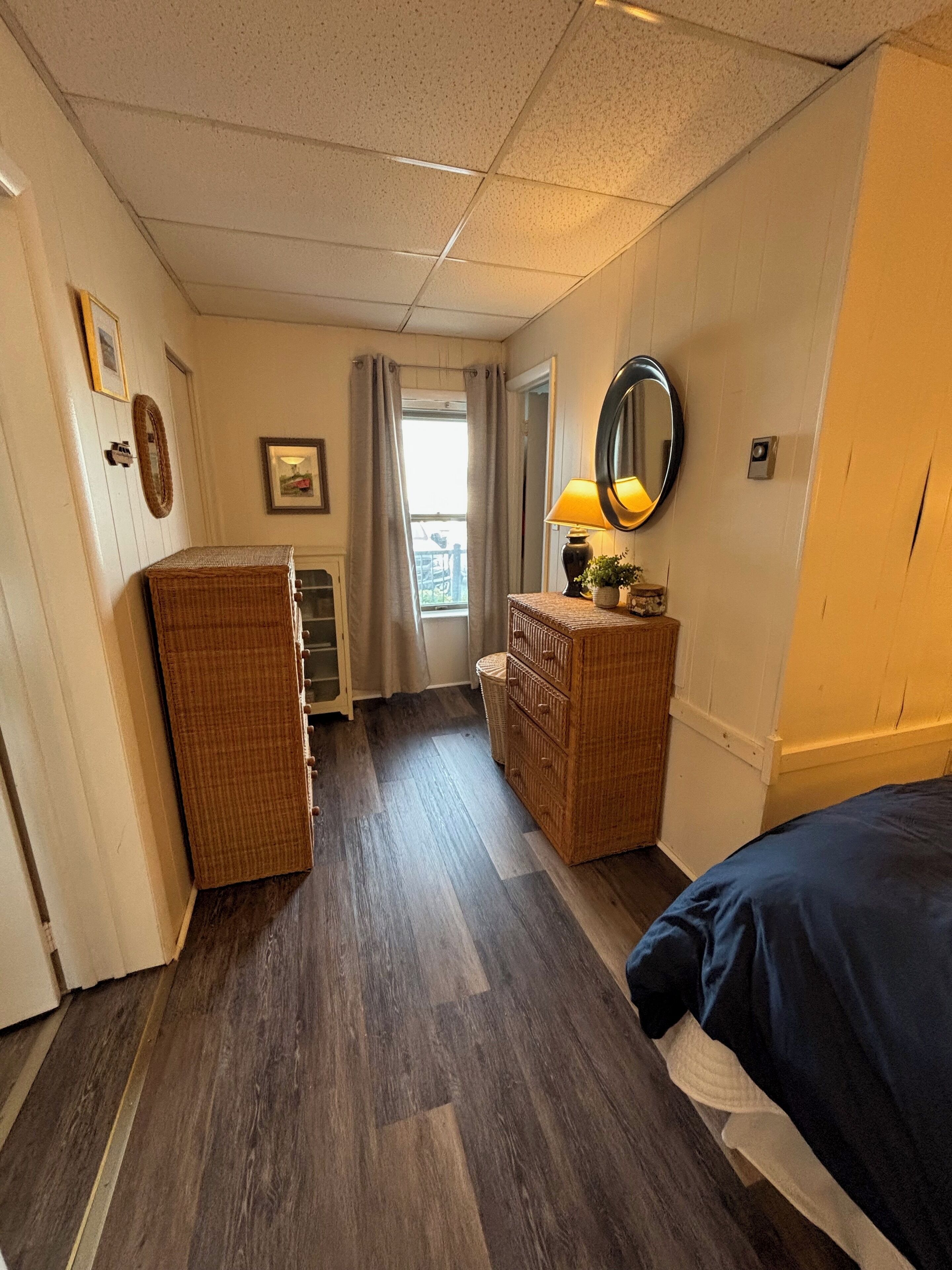 Une chambre d’hôtel avec un plancher en bois, un lit, une commode, un miroir et une fenêtre avec des rideaux.