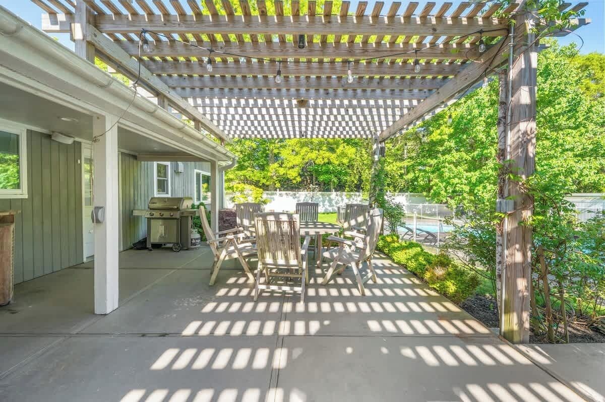 An outdoor patio with a wooden pergola, a dining table with chairs, and a view of a swimming pool and greenery.