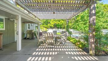 An outdoor patio with a wooden pergola, a dining table with chairs, and a view of a swimming pool and greenery.