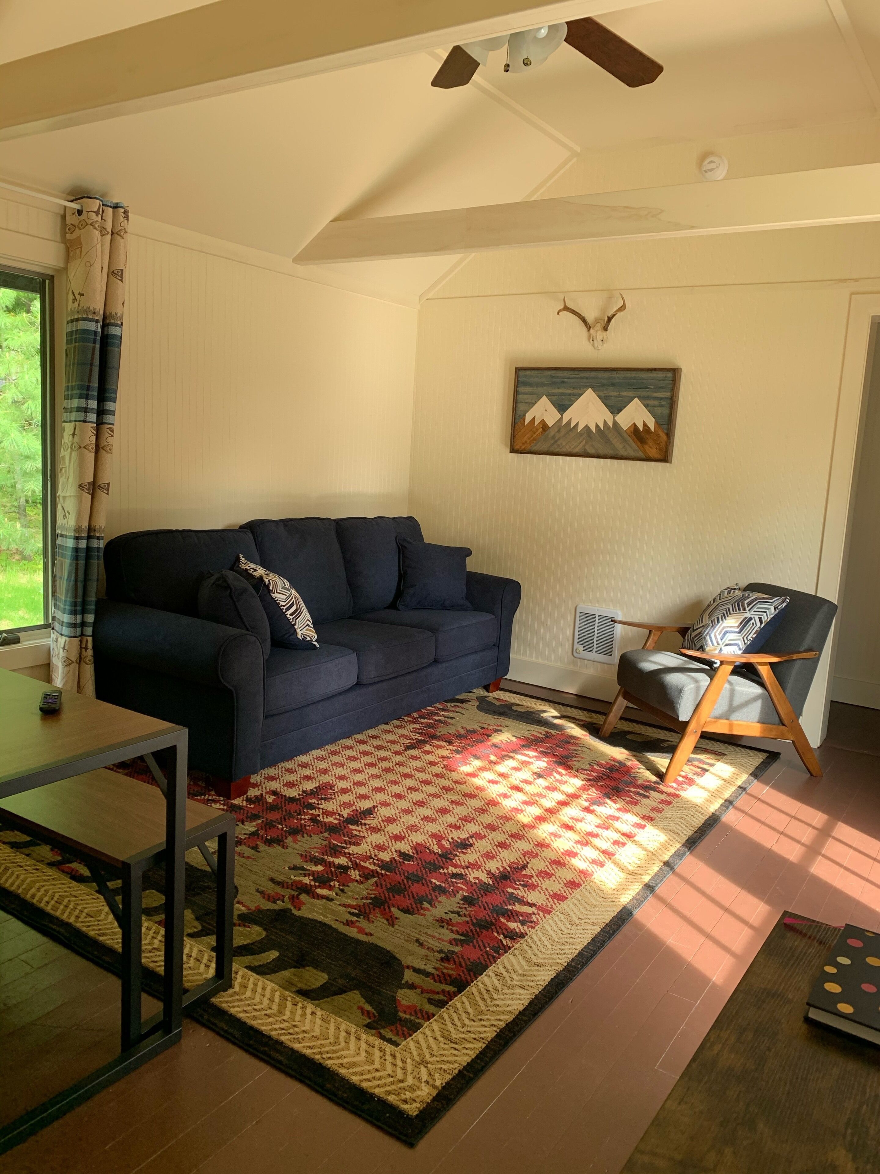 Una sala de estar con un sofá azul oscuro, un tapete estampado, una silla de madera y un cuadro colgado en la pared con un paisaje de montañas.