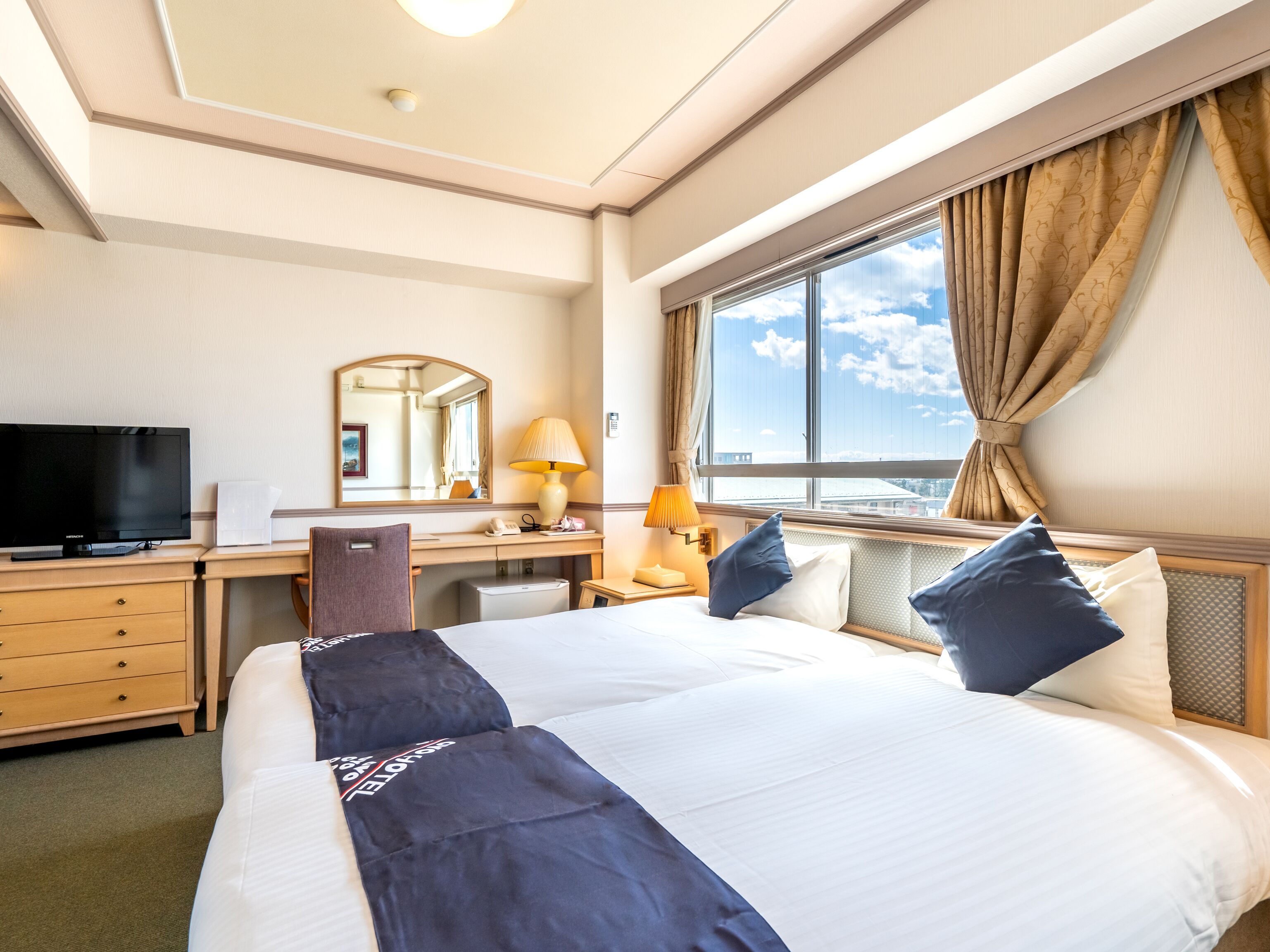 Une chambre d’hôtel avec un grand lit, un bureau avec une télévision, une lampe et une fenêtre avec des rideaux.