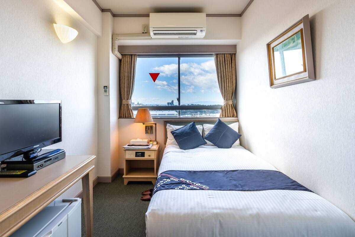 Ein Hotelzimmer mit Bett, Fernseher, Schreibtisch mit Computer, Lampe und Fenster mit Blick auf die Stadt.