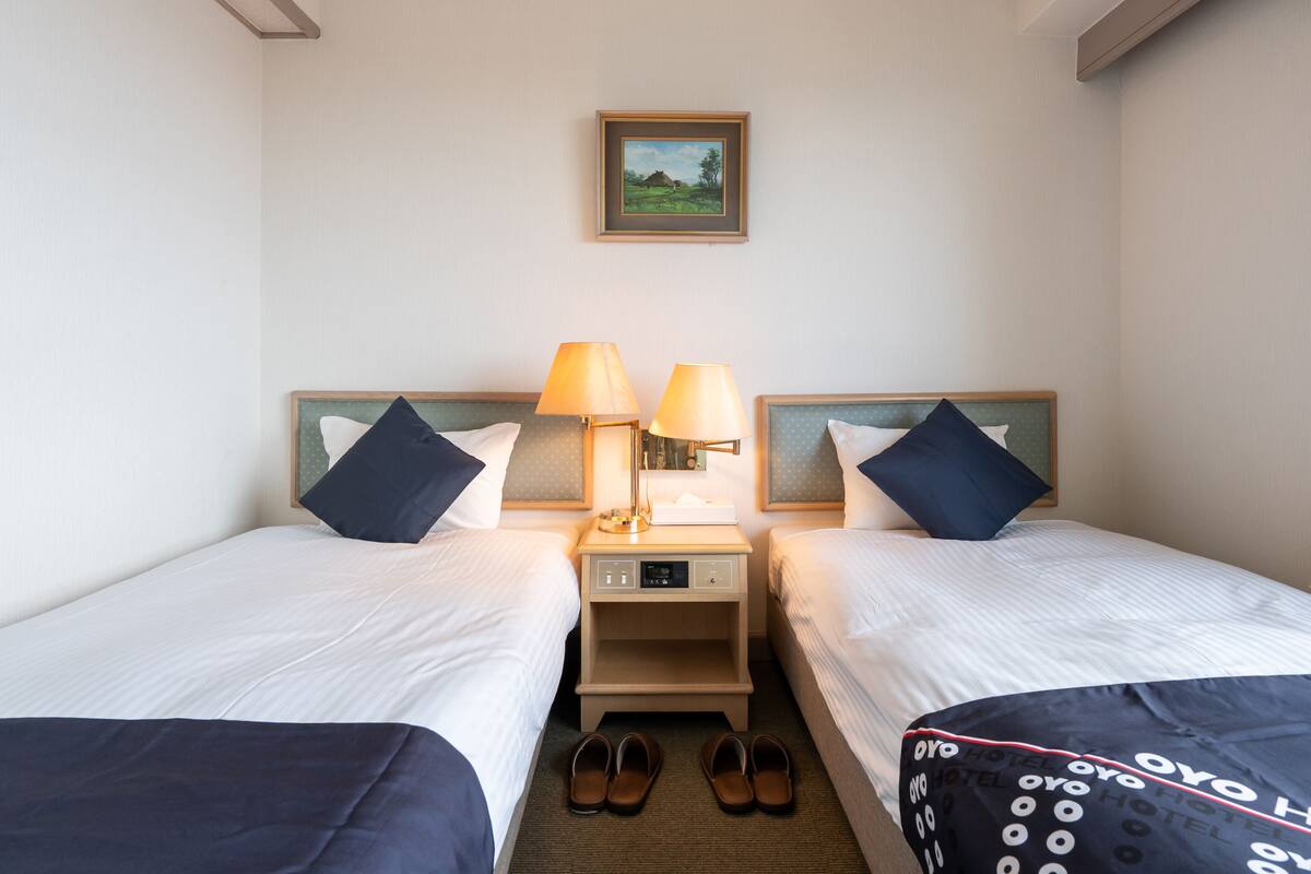 Ein Hotelzimmer mit zwei Betten, einem Nachttisch mit Lampen, einem gerahmten Bild an der Wand und einem Paar Schuhe auf dem Boden.