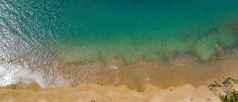 An aerial view of a beach with clear turquoise water, sandy shore, and scattered umbrellas.