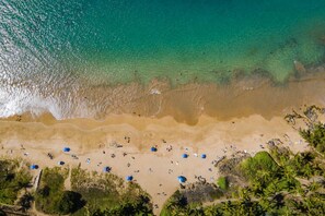 Eine Vogelperspektive eines Strandes mit klarem türkisblauem Wasser, sandigem Ufer und vereinzelten Sonnenschirmen.