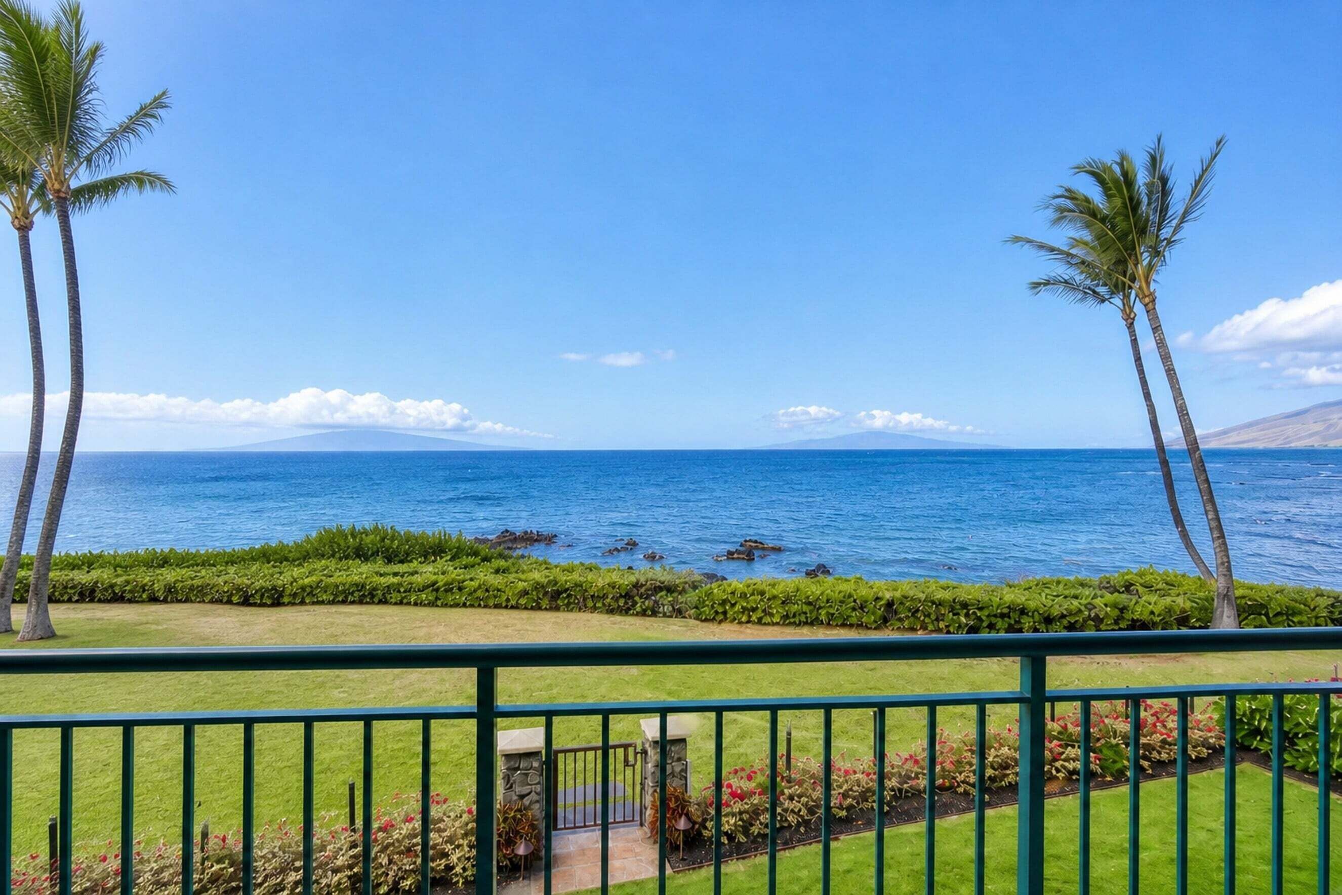 A balcony with a view of the ocean, palm trees, and a well-maintained garden.