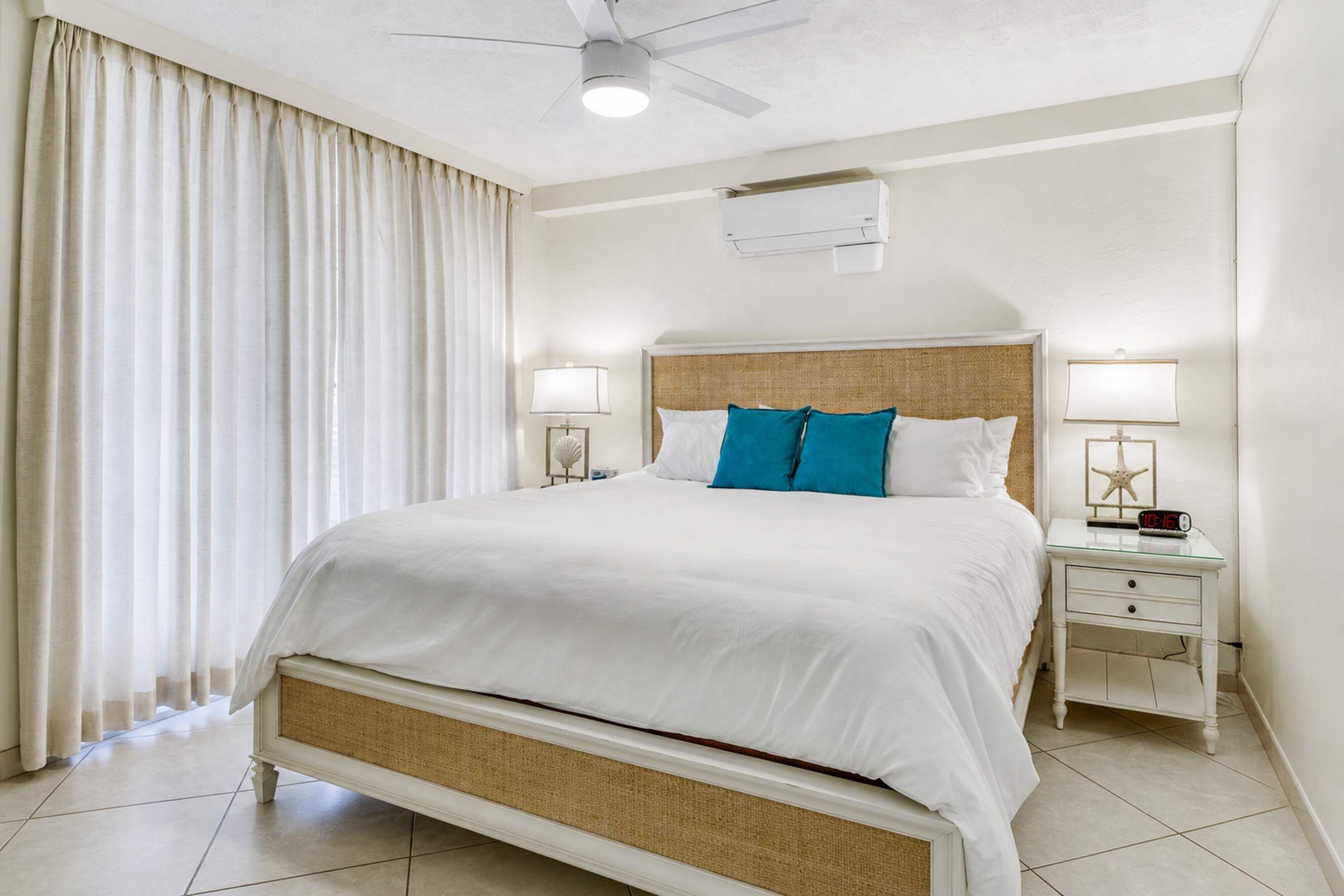 A hotel room with a bed, two bedside tables, a ceiling fan, an air conditioning unit, and a window with curtains.